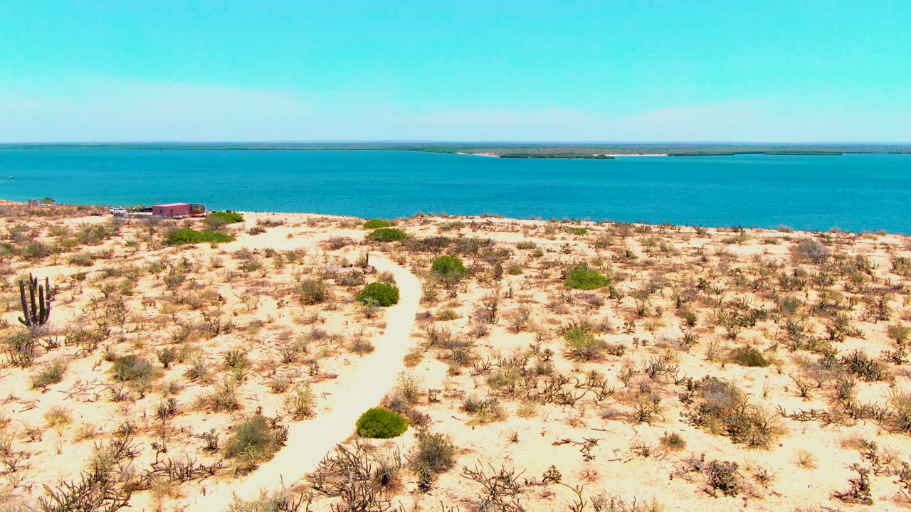 A wide aerial frame shows a faint sandy track through arid vegetation, turquoise water behind the desert, distant low land, and no visible people.