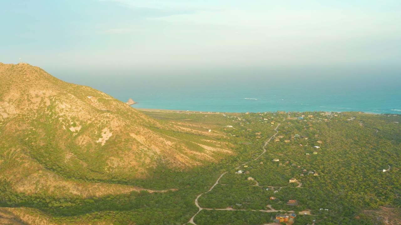 Opening aerial frame over Cabo Pulmo, Baja California Sur, with a rocky mountain on the left, green valley below, a winding road, scattered buildings, blue sea beyond, and no visible people.