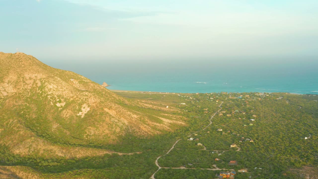 At 0:02, the aerial frame shows the Cabo Pulmo mountain slope left of center, a rural green valley with a visible road and scattered structures, distant turquoise sea, and no visible people.