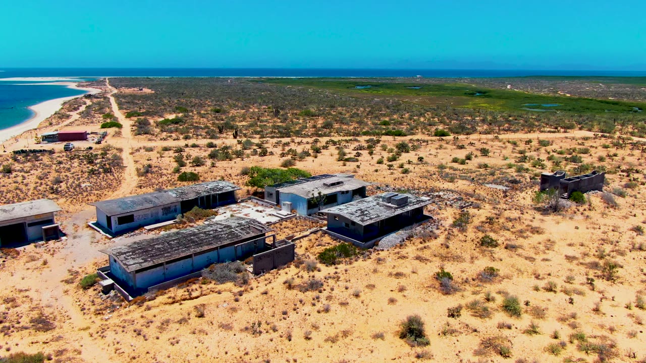 Aerial frame over abandoned flat-roofed buildings in San Carlos, Baja California Sur, with sandy scrubland, a dirt road, distant ocean, clear blue sky, and no visible people.