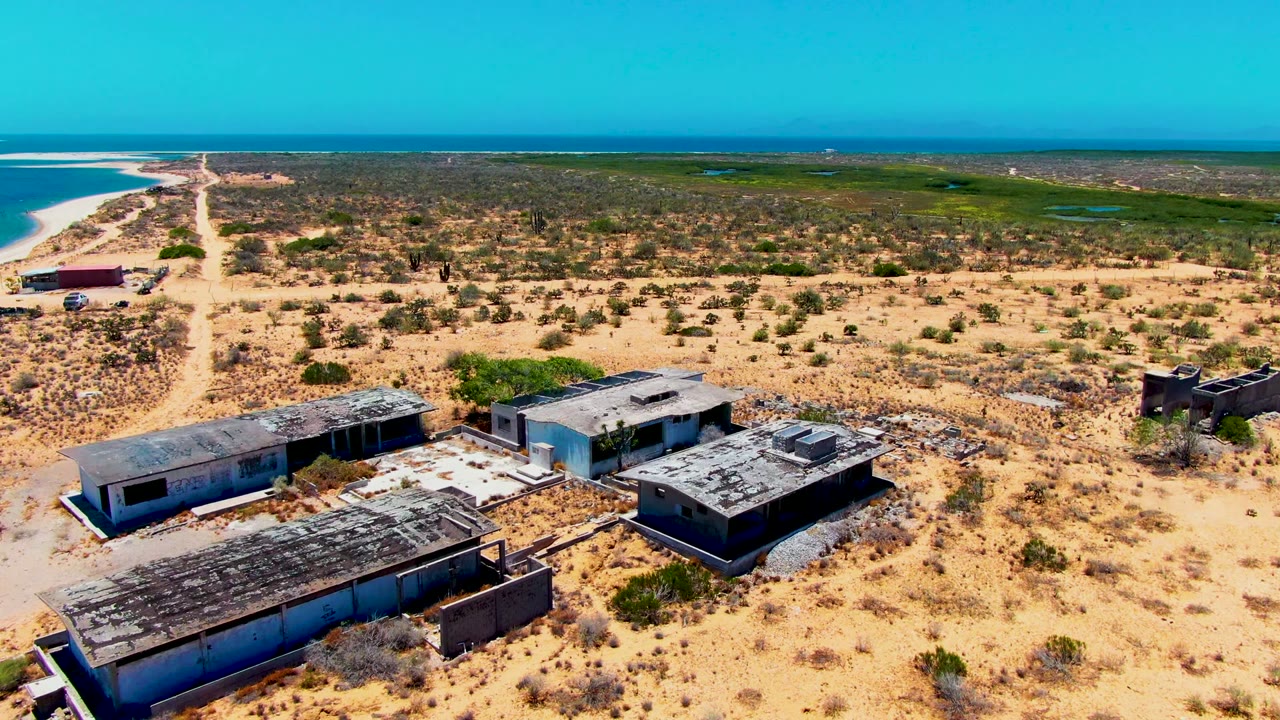 Drone view of dilapidated buildings beside sandy scrub and a distant shoreline.