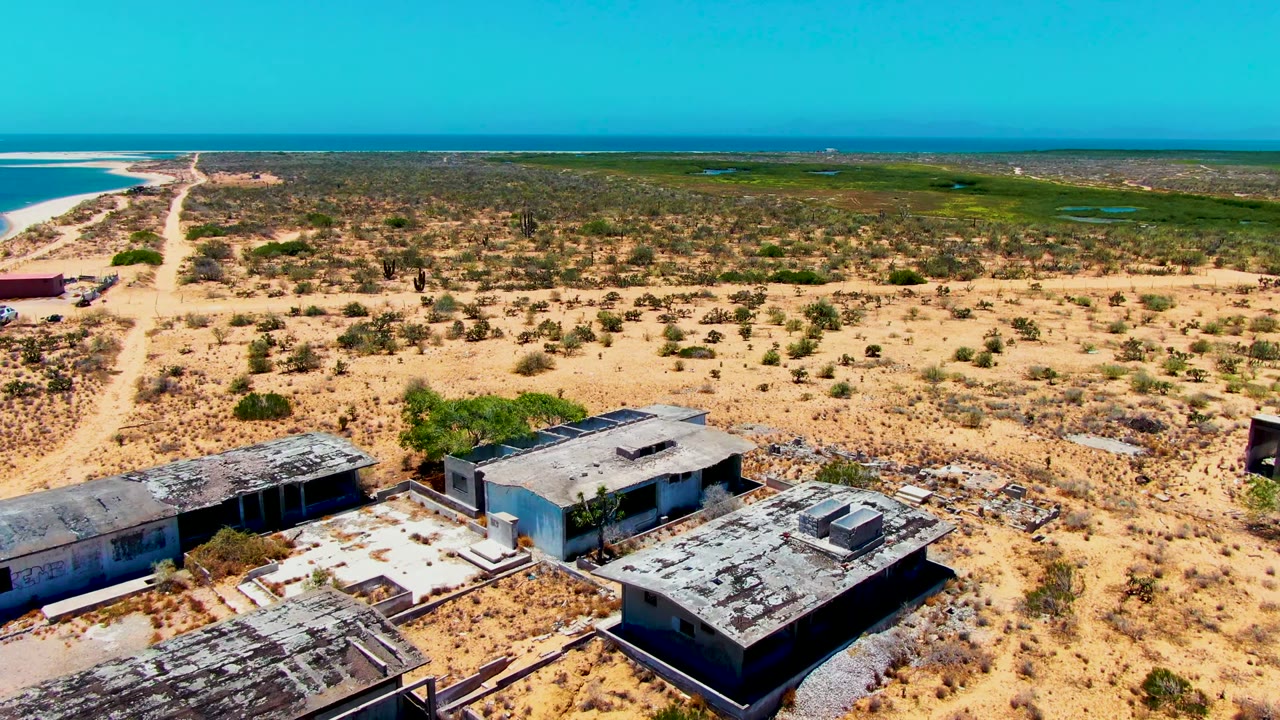 High aerial frame over roofless and weathered structures in San Carlos, Baja California Sur, with open desert vegetation, a distant ocean line, blue sky, and no visible people.