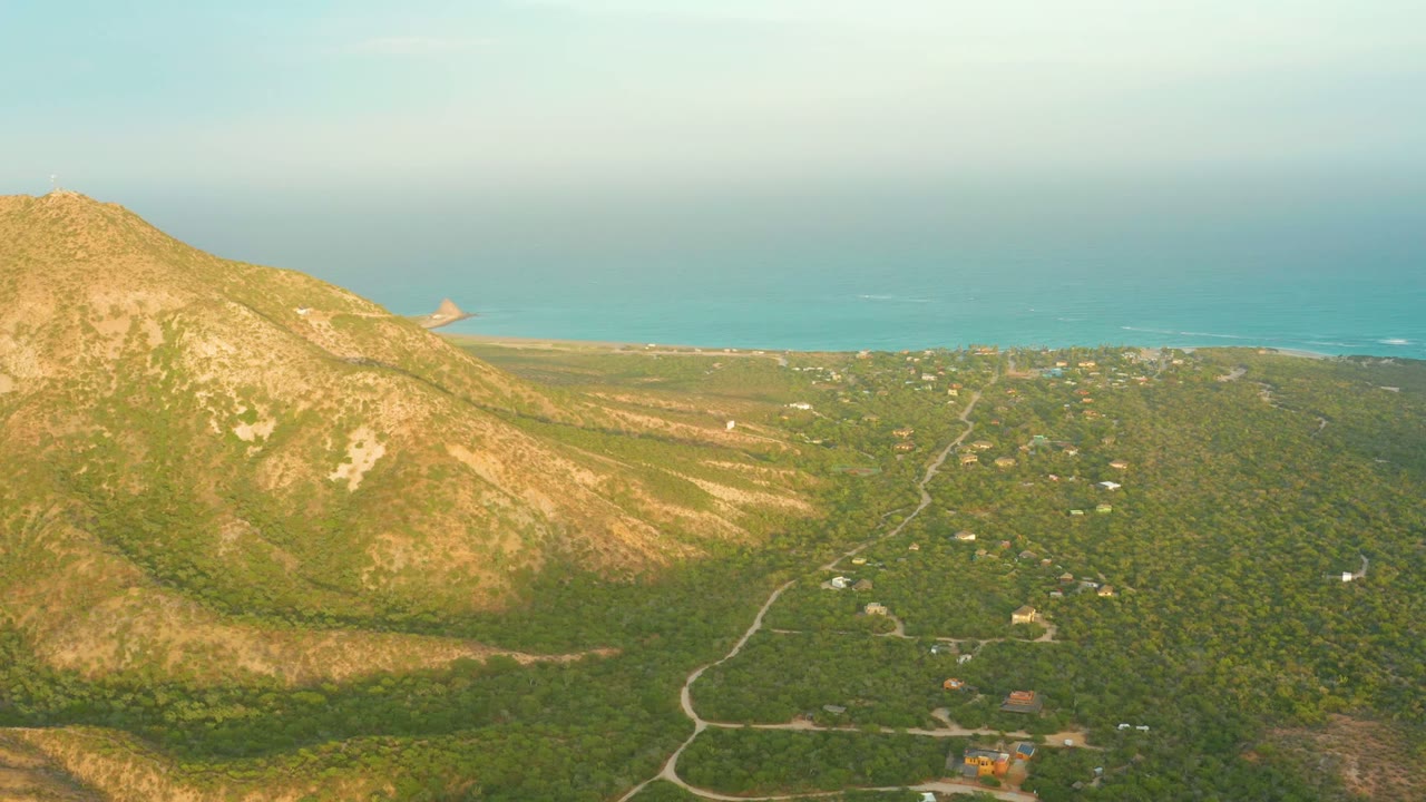 Opening aerial frame over Cabo Pulmo with a sunlit rocky mountain on the left, green vegetation below, dirt roads through the valley, blue ocean on the horizon, and no visible people.