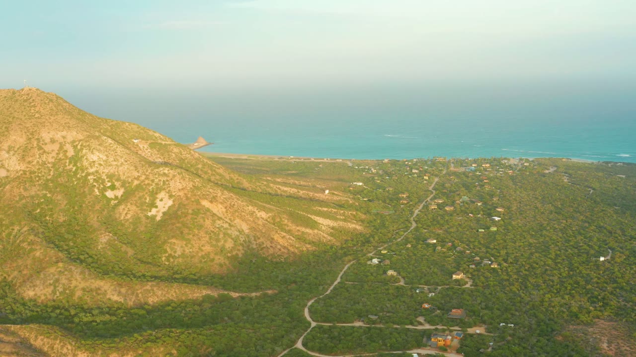 Wide aerial landscape with mountain slope, green scrubland, roads, scattered buildings, and sea.