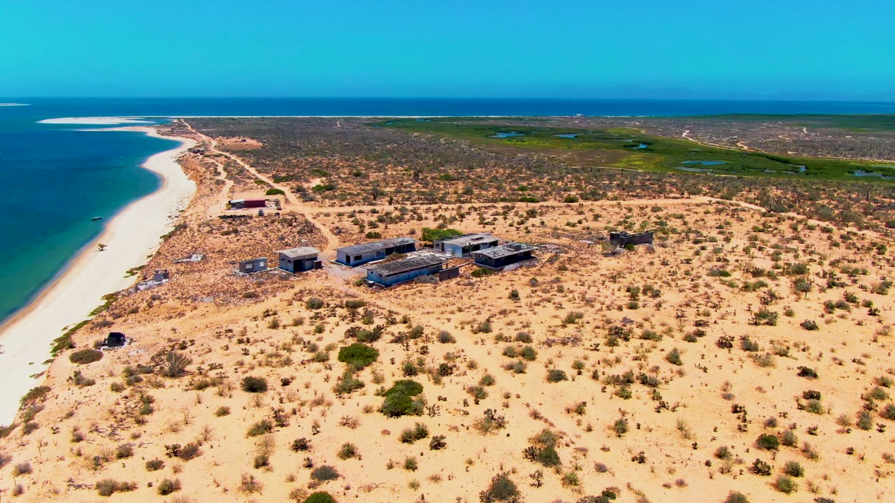 Drone view of ocean shoreline beside arid sand and a cluster of low buildings.