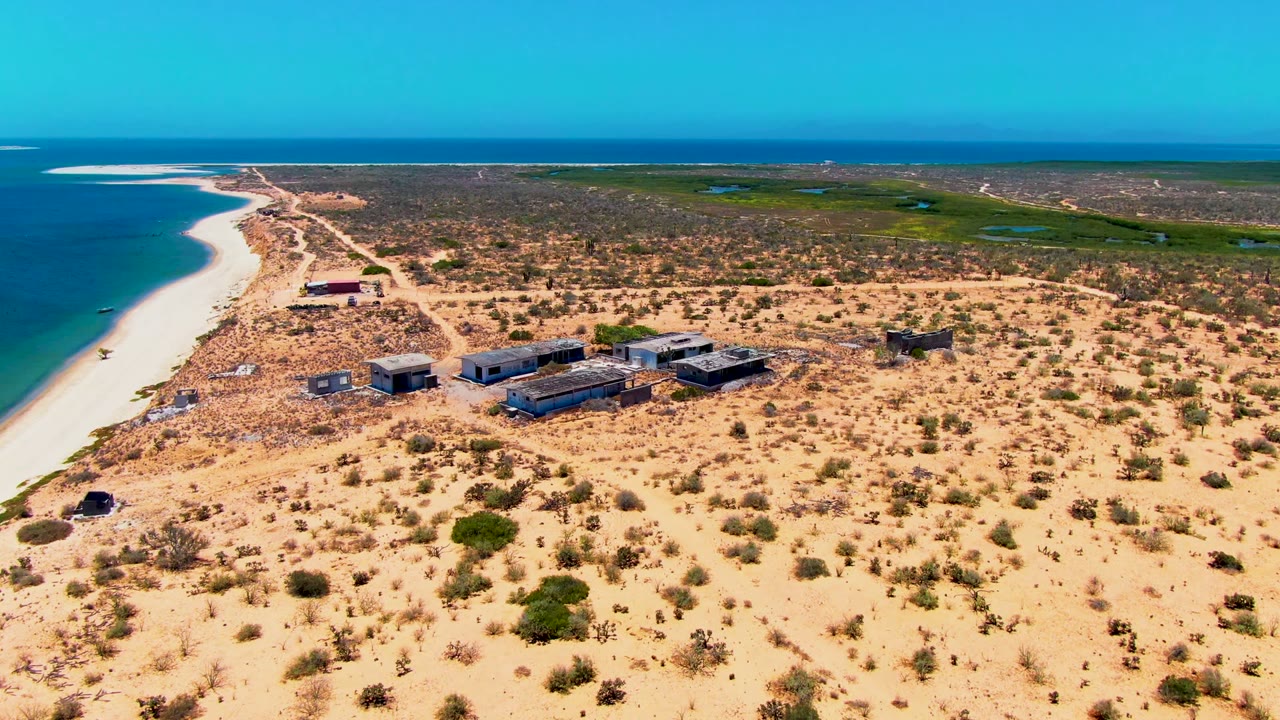Aerial frame from San Carlos, Baja California Sur with blue ocean, a narrow pale beach, a sandy track, low structures near shore, scattered shrubs inland, and no visible people.