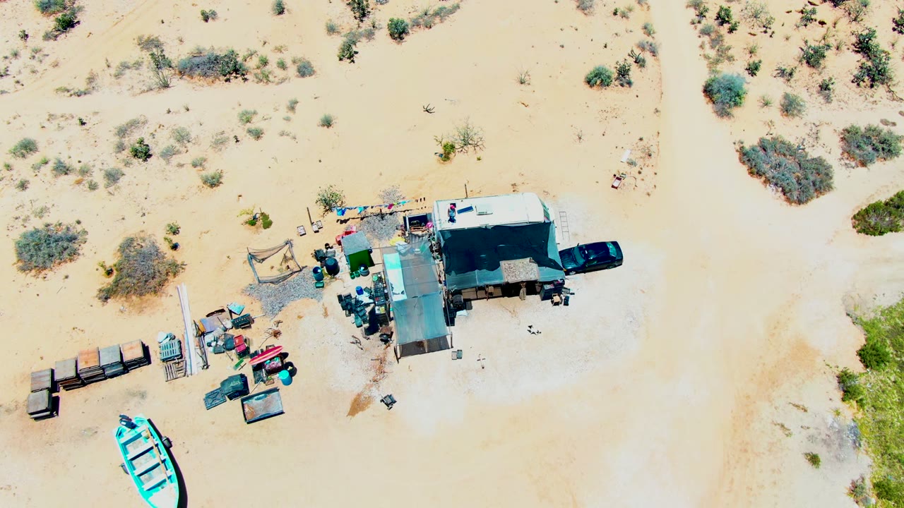 Top-down view of a desert camp in San Carlos, Baja California Sur, with a white trailer, black car, scattered gear, scrub vegetation, and no visible people.