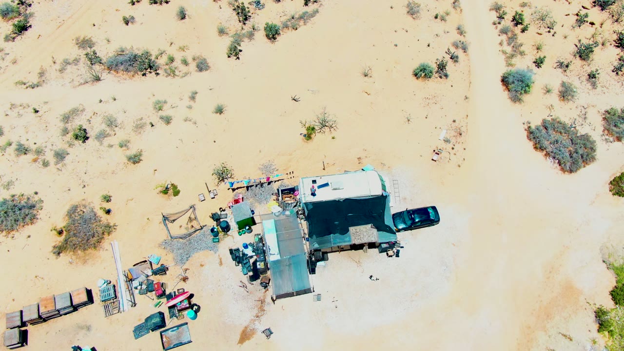 Overhead San Carlos campsite frame with the white trailer, black car, shade structure, small containers, and patchy desert brush visible against pale sand, no visible people.