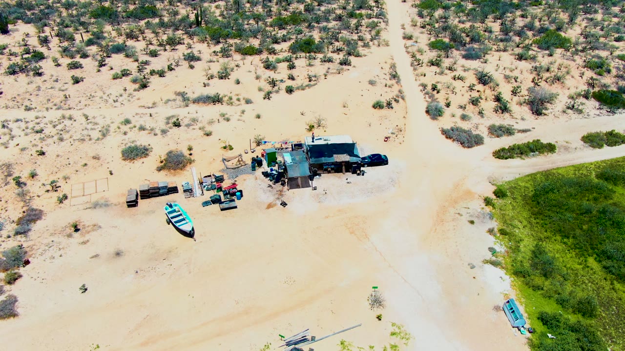Overhead drone frame in San Carlos, Baja California Sur showing a small structure, parked vehicles, trailers or gear, pale sand, sparse scrub, a dirt road, a green patch, and a blue boat, with no visible people.