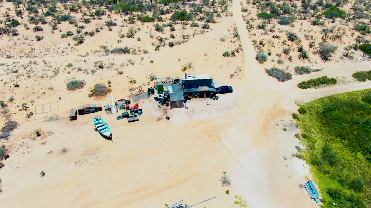 Aerial frame in San Carlos, Baja California Sur with the small desert structure near the top, clustered machinery and gear beside it, open sand through the center, and the blue boat lower in frame, with no visible people.