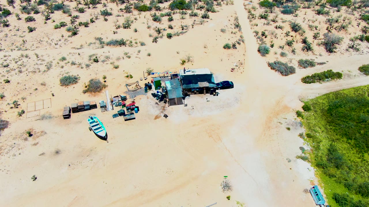 Overhead frame in San Carlos, Baja California Sur where the dirt road runs down the middle, scrub fills the upper desert, the structure and vehicles sit to the right, and the blue boat is near the green patch, with no visible people.