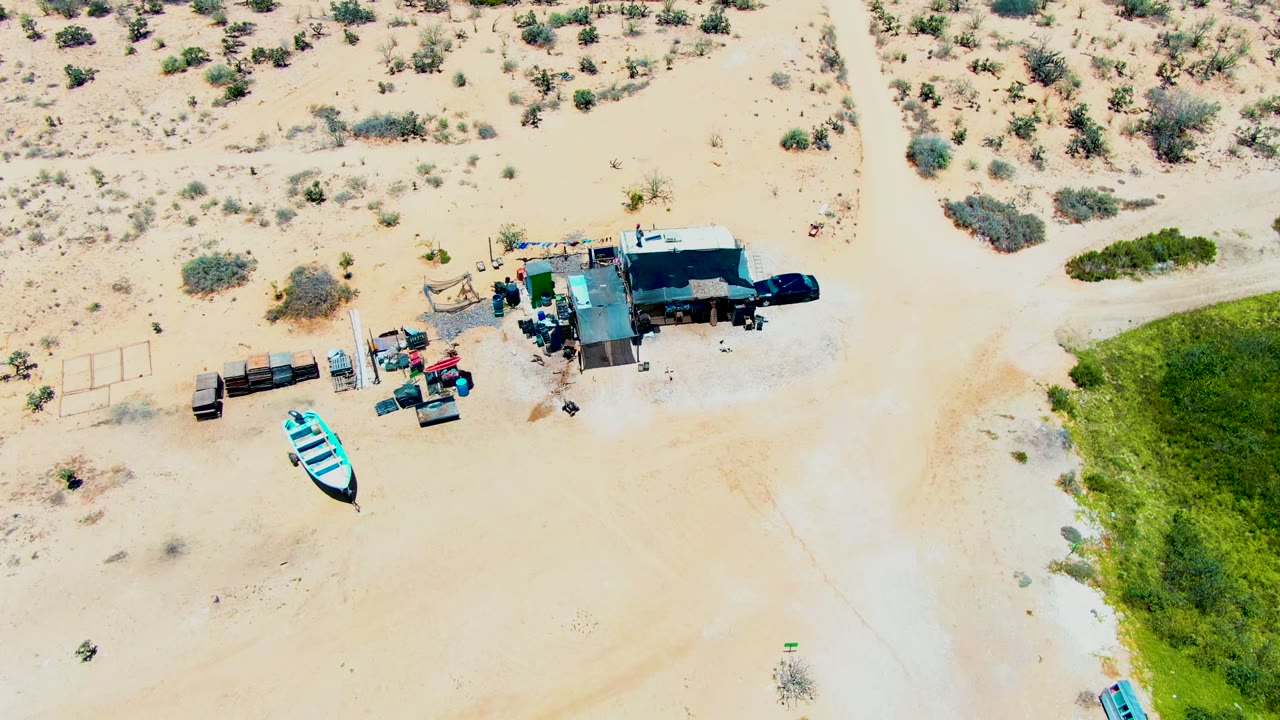 High-angle desert campsite with trailers, machinery, a truck, and a blue boat on sand.