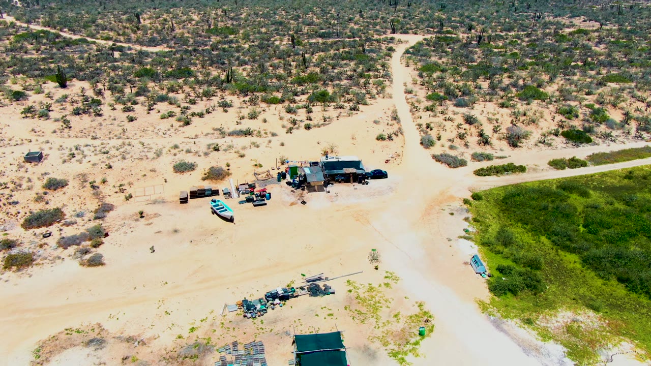 Opening overhead view in San Carlos, Baja California Sur, with pale sandy tracks, sparse shrubs, a blue boat near a small structure, a bright green field at right, and no visible people.