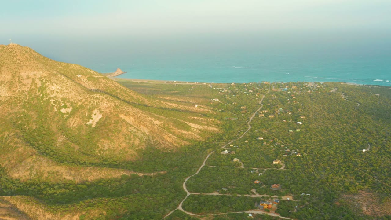 Aerial view in Cabo Pulmo, Baja California Sur, Mexico, with a sunlit mountain on the left, green valley roads below, scattered buildings, blue sea on the horizon, and no visible people.