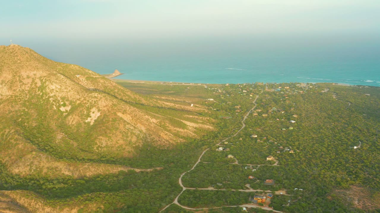 Cabo Pulmo aerial view with the mountain on the left edge, dense green valley terrain across the frame, thin roads, scattered structures, blue water, and no visible people.