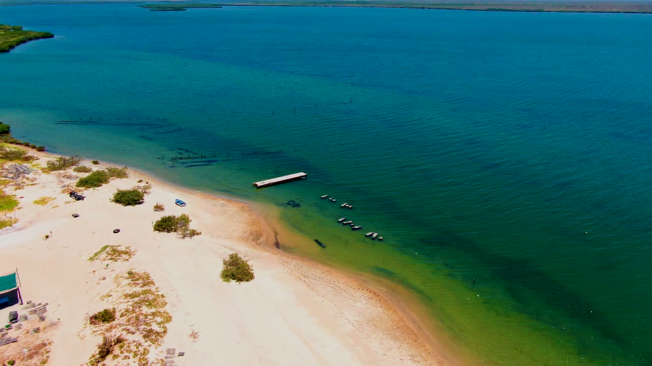 Aerial frame over San Carlos, Baja California Sur showing pale sand, scattered shrubs, a dock in shallow turquoise water, small boats nearby, and no visible people.