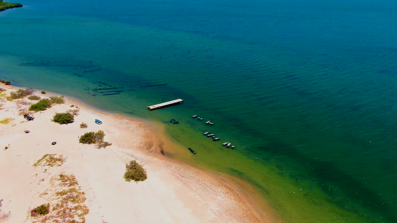 San Carlos, Baja California Sur beach seen from above, with bright sand, low vegetation, a small dock, several boats in shallow water, darker offshore lines, and no visible people.