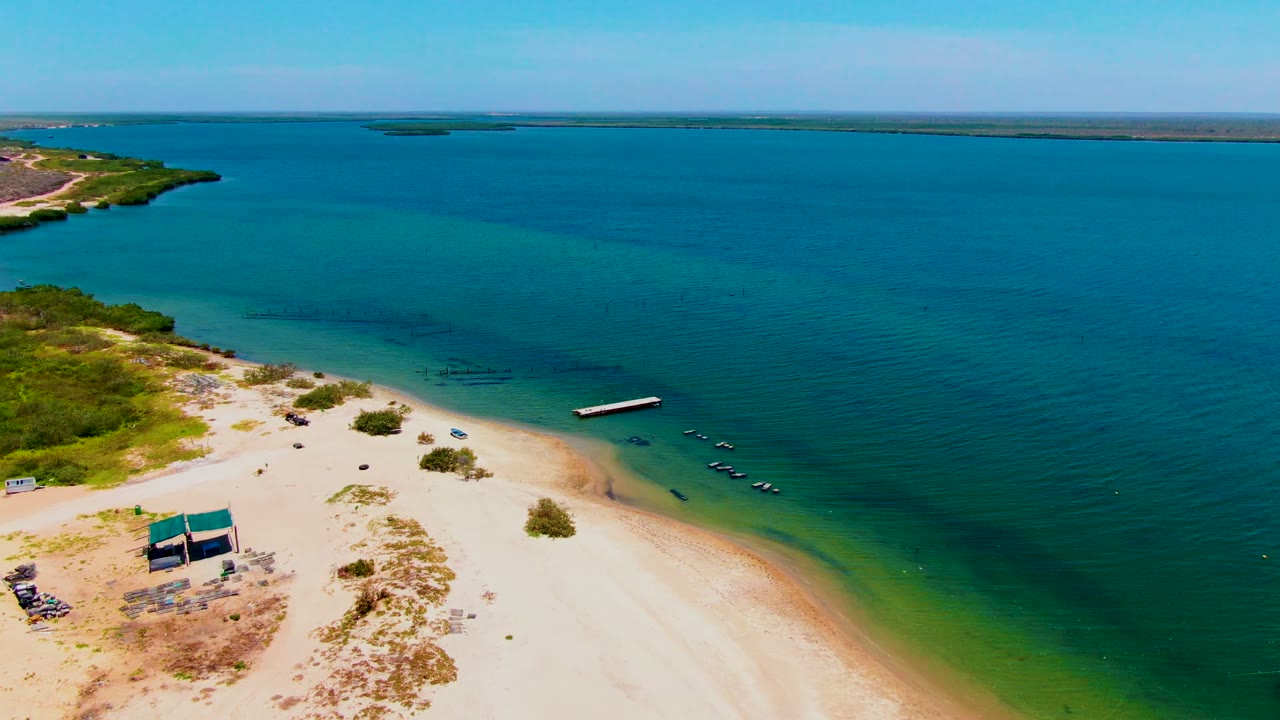 San Carlos aerial frame showing the beach shelter at lower left, pale sand across the foreground, clear coastal water to the right, and no visible people.