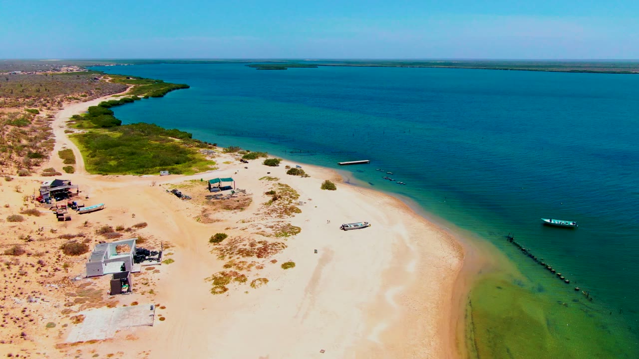 Aerial frame over San Carlos, Baja California Sur showing pale sand, turquoise shallows, small boats, low beach structures, scrub greenery, and no visible people at the start of the clip.