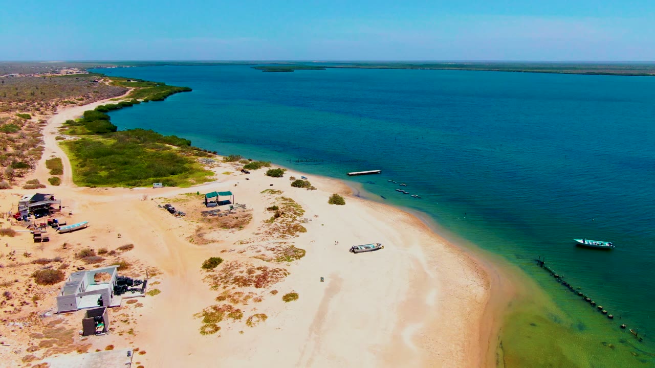 Drone view along a sandy beach with calm blue water, small boats, and sparse structures.