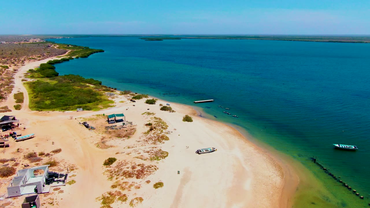 Aerial frame over San Carlos, Baja California Sur showing more open blue water, the sandy beach at lower left, small boats near shore, beach shelters, green brush, and no visible people.