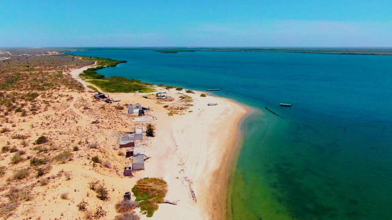 Aerial frame over San Carlos, Baja California Sur, showing pale sand, several small shoreline structures, sparse desert scrub, calm blue-green water, anchored boats, and no visible people.