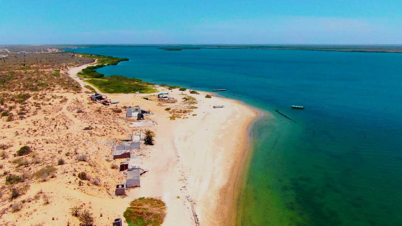 Elevated coastal view with huts near the sand, boats offshore, and shallow green water along the beach.