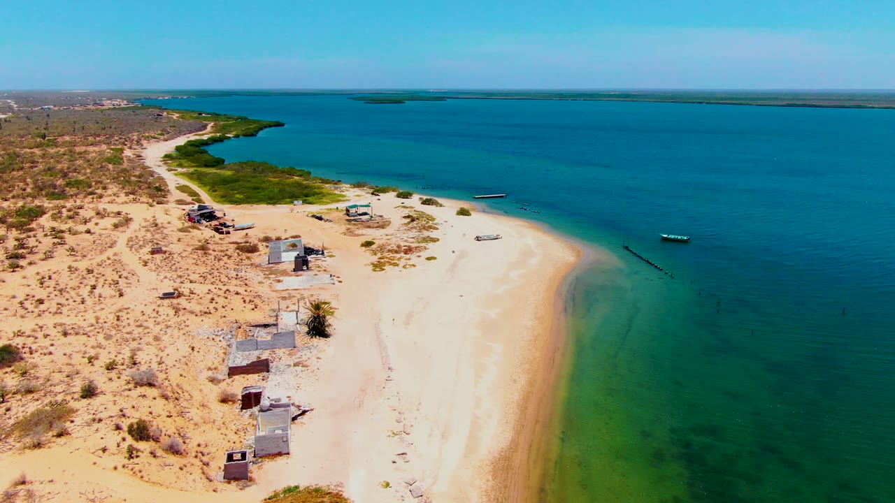 Drone-height frame over San Carlos, Baja California Sur, with a sandy coastal point, scattered small structures, scrubby desert ground, several boats offshore, and no visible people.