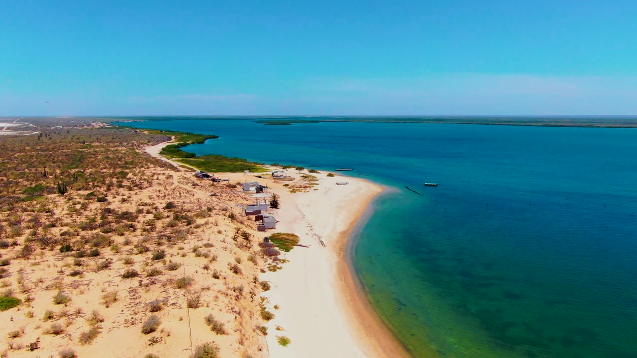 Opening aerial frame over San Carlos, Baja California Sur, showing pale sand curving around blue-green water, dry scrubland inland, small huts near the shore, distant boats, and no visible people.