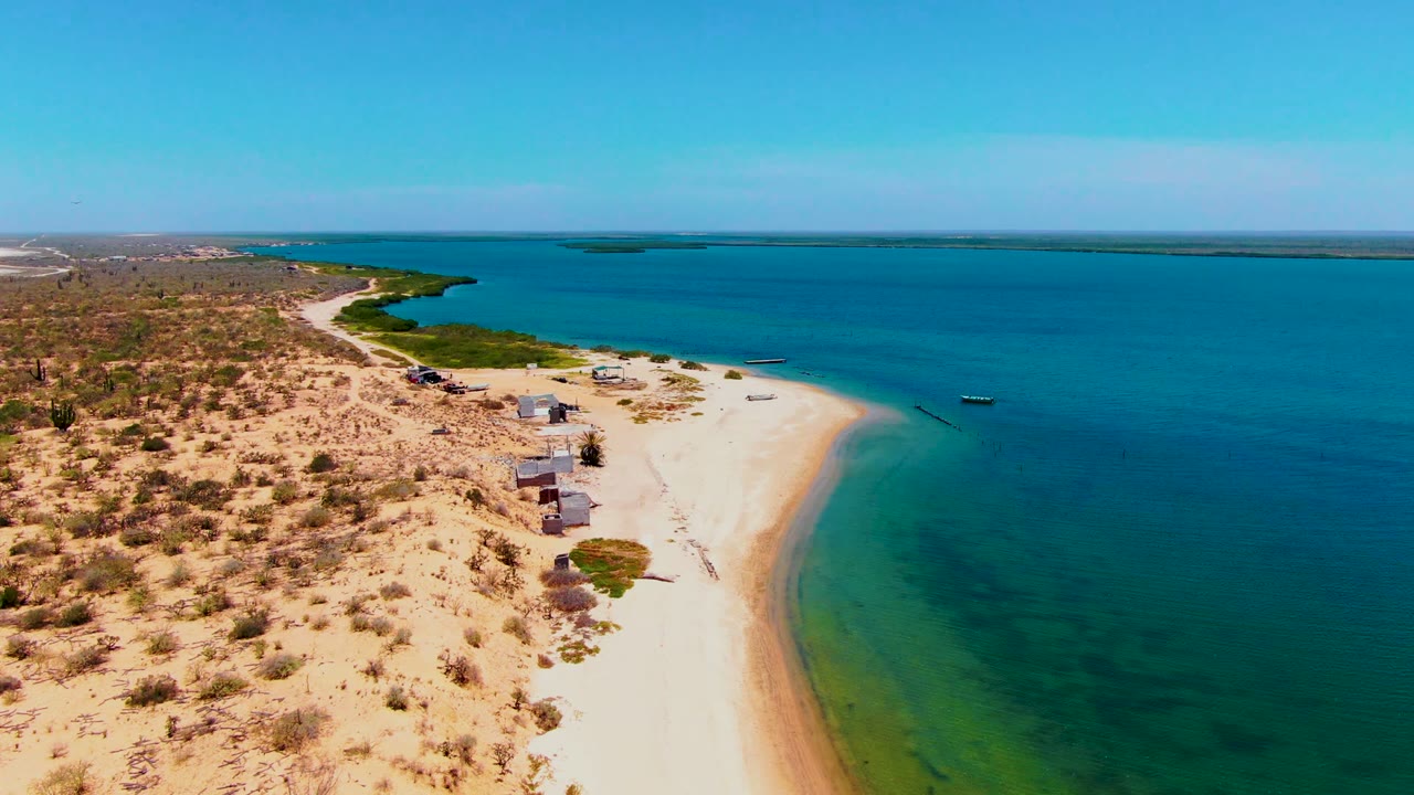 San Carlos coastal frame showing the sandy point sweeping into blue water, small huts along the beach, sparse desert vegetation to the left, and no visible people.