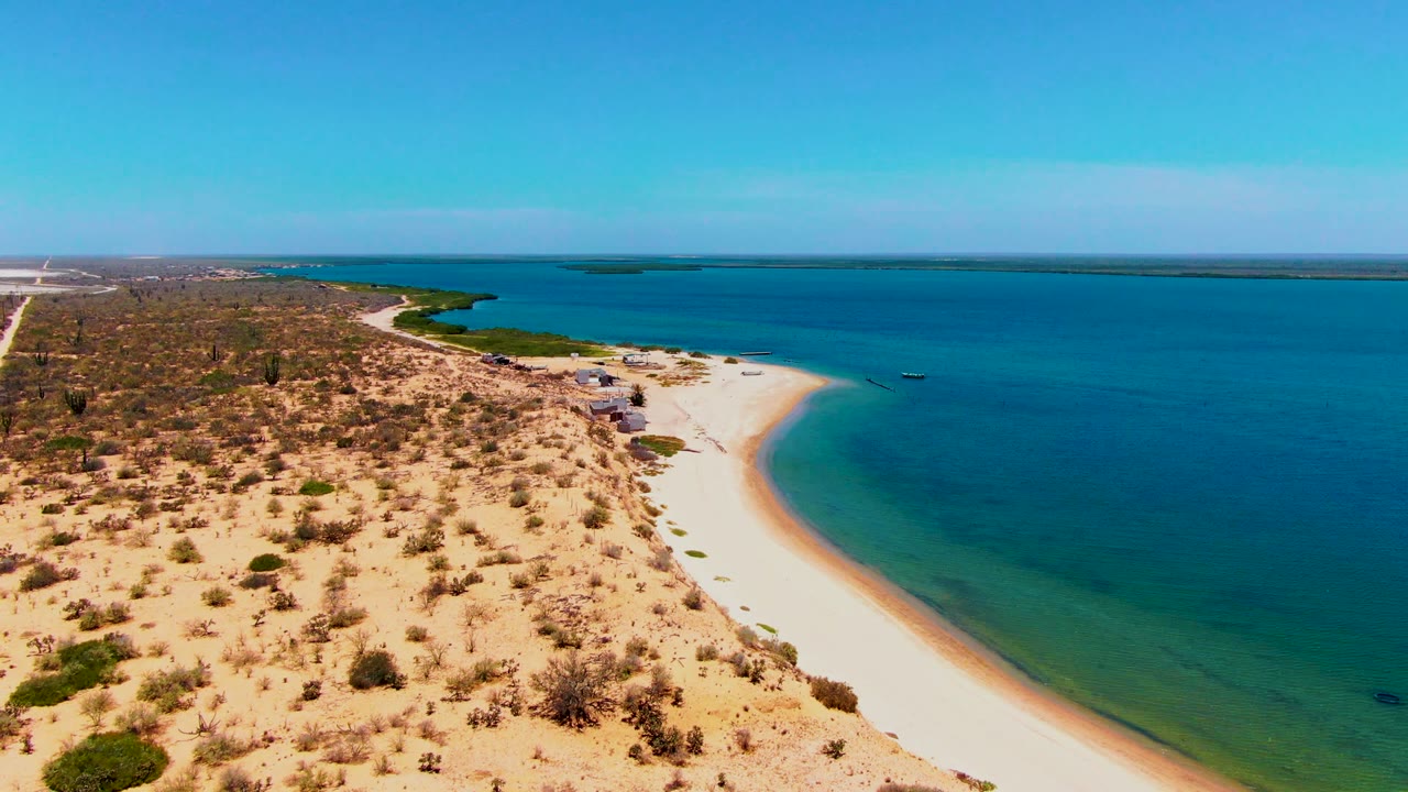 Aerial frame over San Carlos, Baja California Sur, showing a broad strip of sandy beach between dry shrub-covered land and blue-green ocean, with low structures near the shore and no visible people.