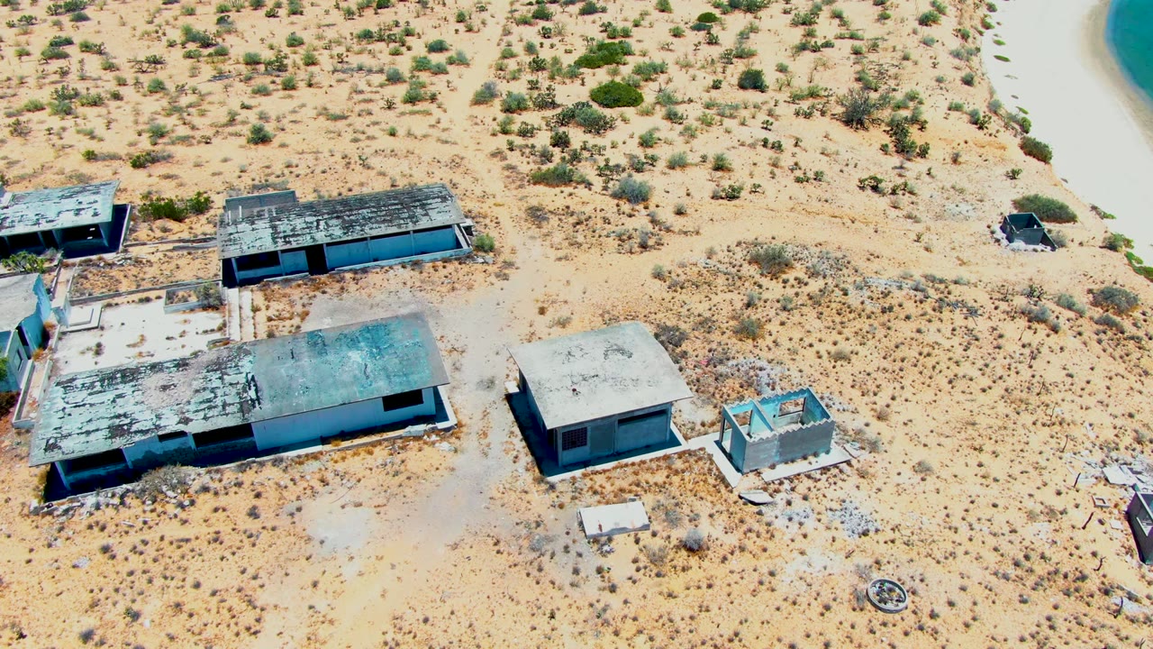 High aerial frame over abandoned concrete structures in San Carlos, Baja California Sur, with sandy ground, scattered shrubs, and no visible people.