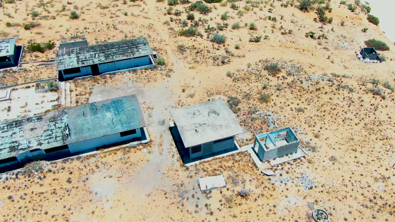 Drone-height frame over weathered buildings in San Carlos, Baja California Sur, with dark roof patches, beige sand, low shrubs, and no visible people.