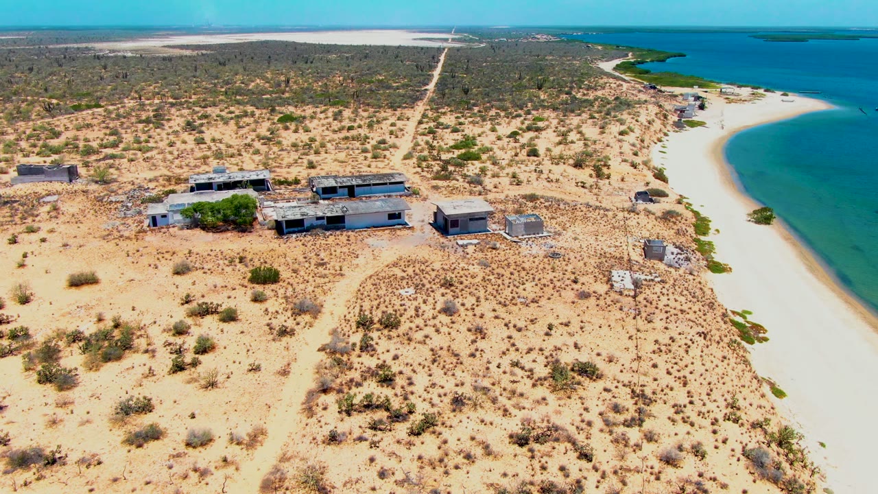 Opening aerial frame over San Carlos, Baja California Sur, with low buildings on sandy scrubland beside a white beach and blue water, no visible people.