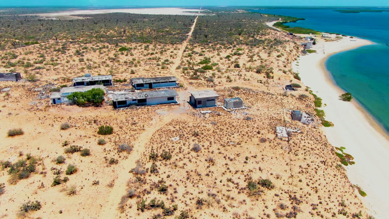Drone view closer to low buildings on arid sand beside a curved beach.