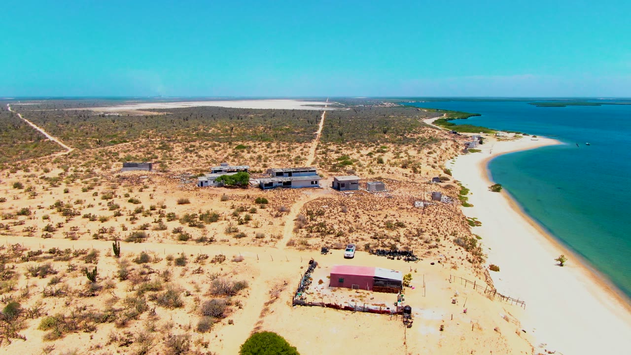 Aerial frame over San Carlos, Baja California Sur, with sparse desert vegetation, scattered buildings, a dirt road, a white vehicle, beach, blue water, and no visible people.