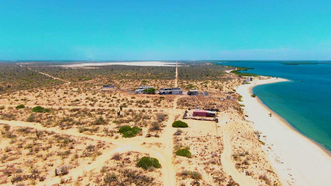 Aerial frame over San Carlos, Baja California Sur showing a diagonal sandy road through desert scrub, clustered low buildings near the shore, bright beach sand, blue coastal water, and no visible people.