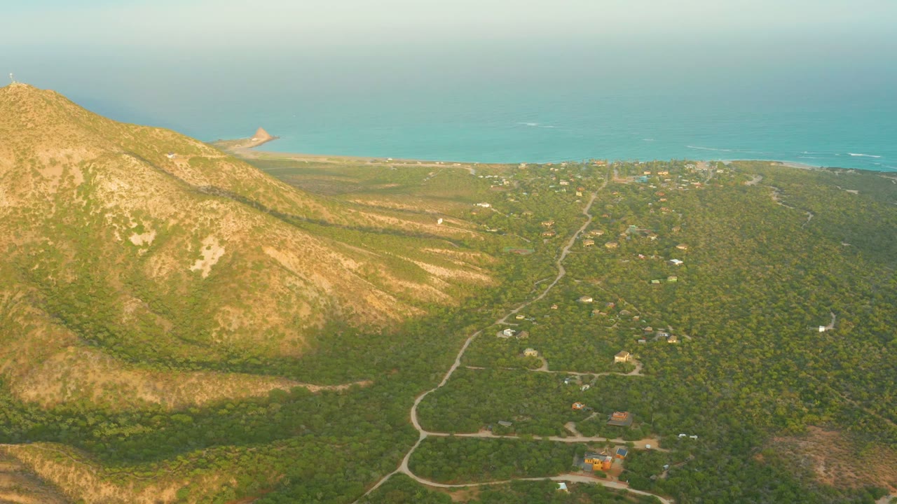 Aerial frame over Cabo Pulmo, Baja California Sur, with a sunlit rocky mountain on the left, green forest below, winding roads, distant coastal water, and no visible people.