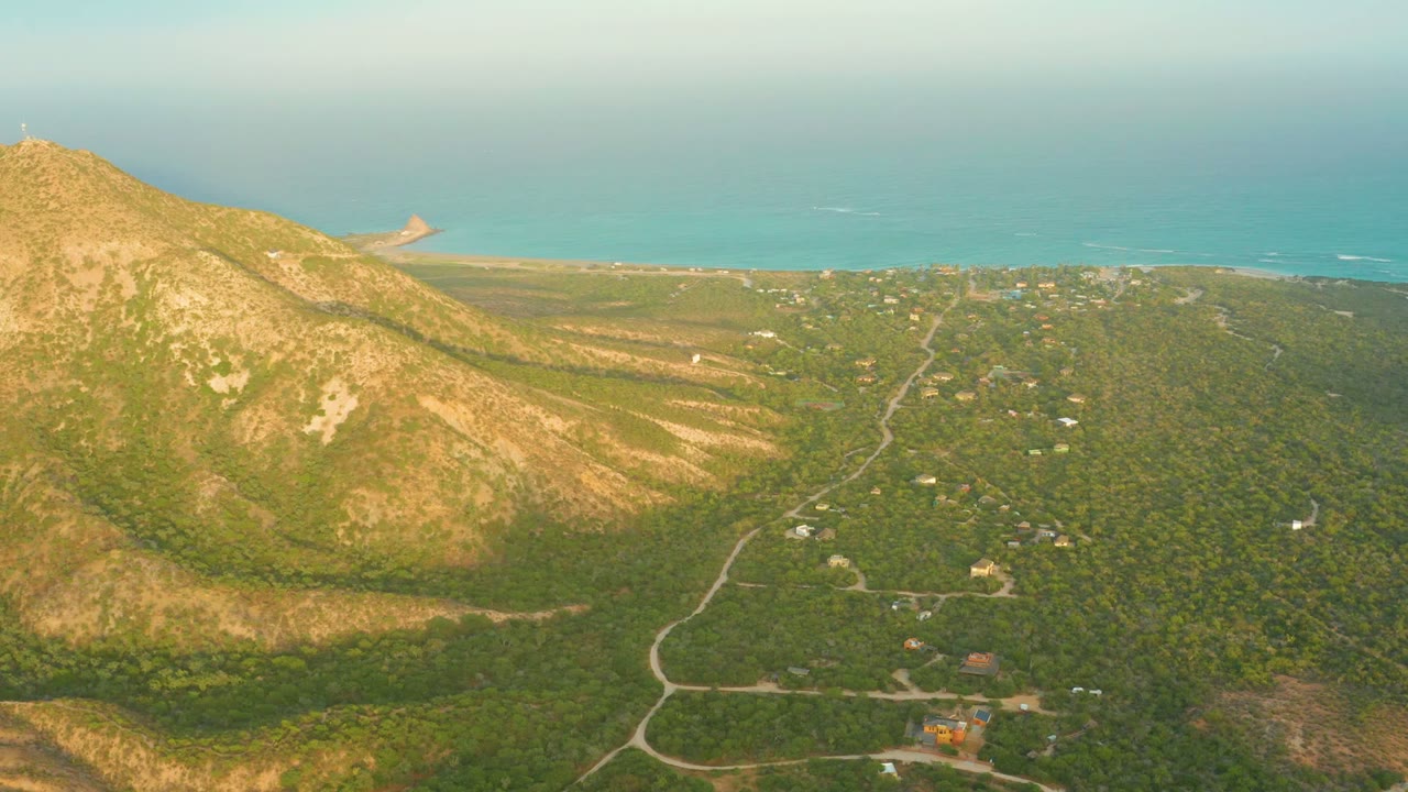 Wide Cabo Pulmo frame showing warm light across the mountain face, dense green terrain to the right, a road running toward the coast, distant structures, and no visible people.