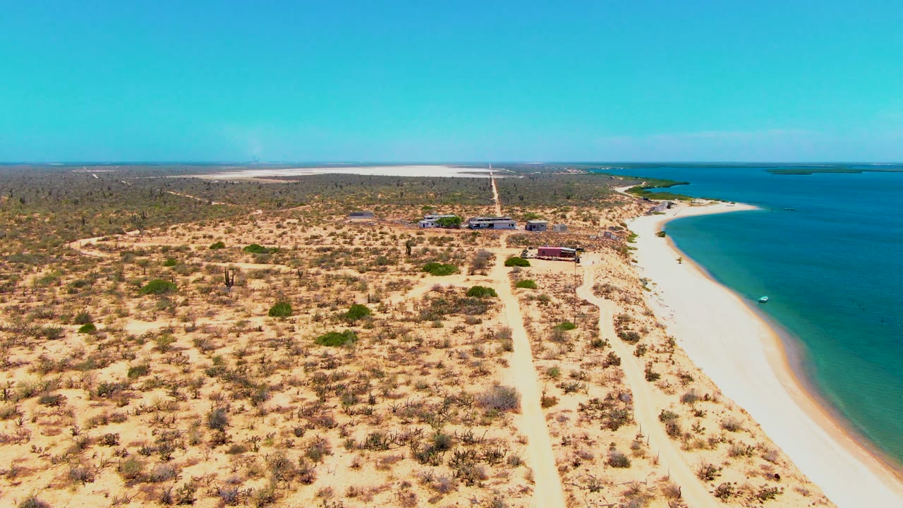 Aerial frame over San Carlos, Baja California Sur showing sandy dunes in front, a straight road through scrub, beachside structures, turquoise ocean, and no clearly visible person at this moment.