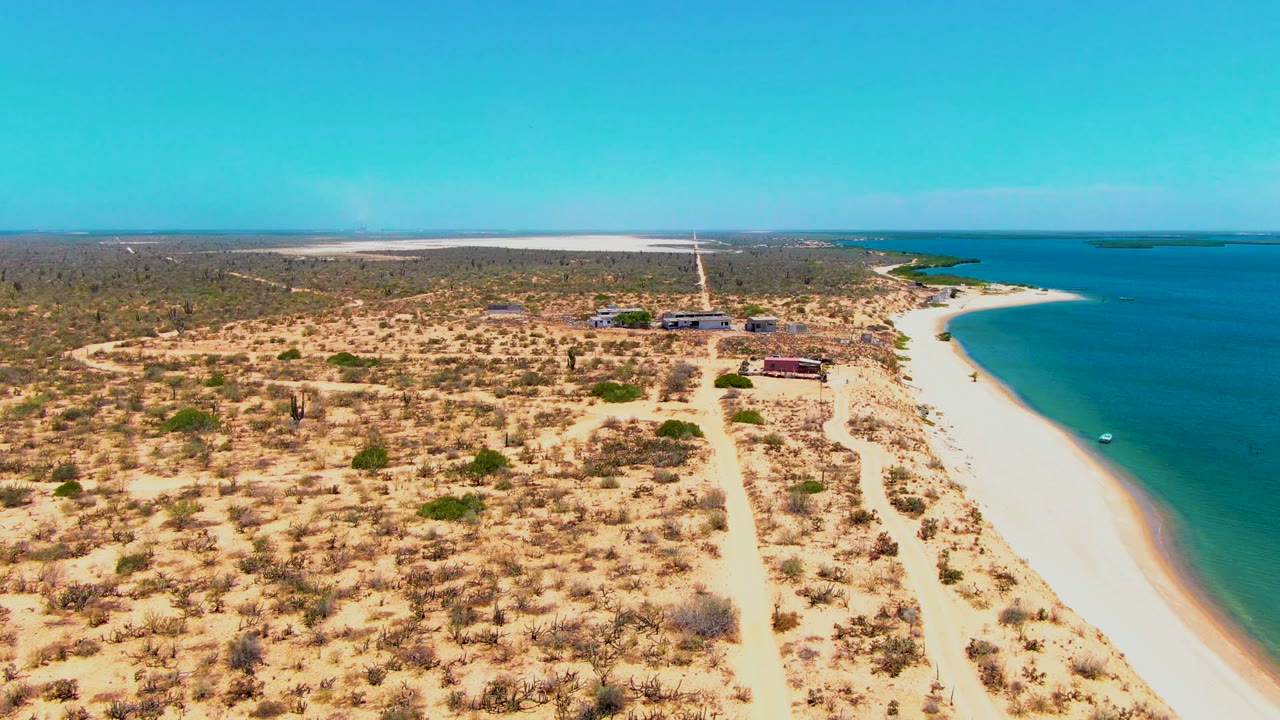 Drone view of coastal dunes, pale beach, turquoise water, and inland road.