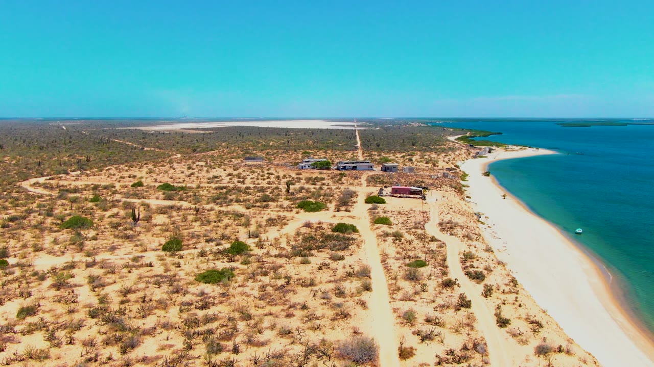The San Carlos coastline fills the frame with dry dunes and scrub on the left, a beach road running toward the horizon, small structures near the sand, and blue-green ocean to the right.