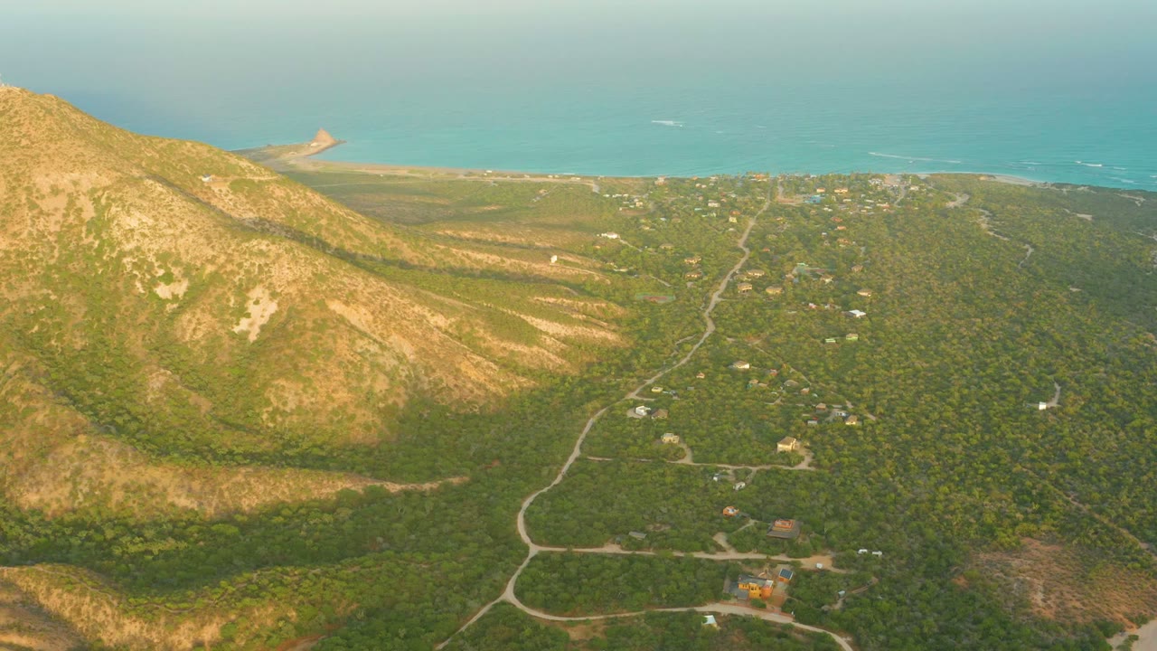 Opening aerial frame over Cabo Pulmo Los Cabos shows a tan-green mountain slope at left, winding dirt roads through brush, scattered buildings, a narrow beach, blue ocean, and no visible people.