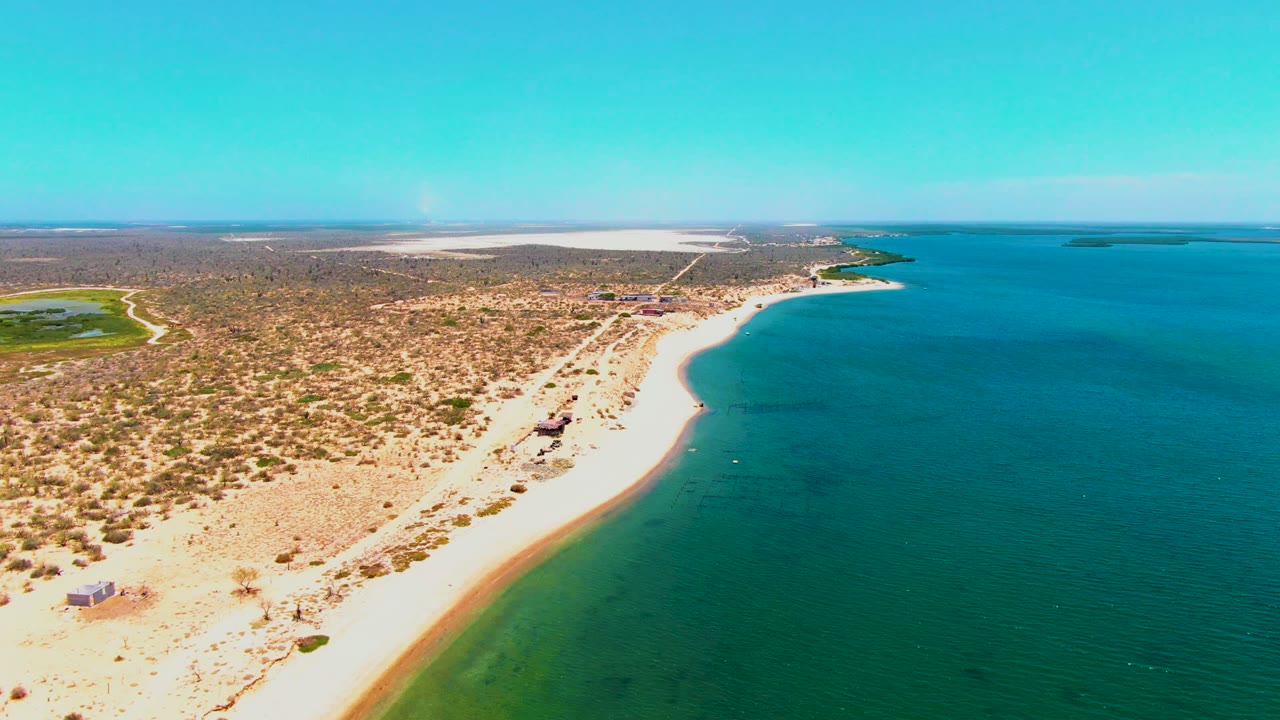 Aerial view over San Carlos, Baja California Sur, with a pale sandy beach curving beside blue water, dry scrubland inland, and no visible people.