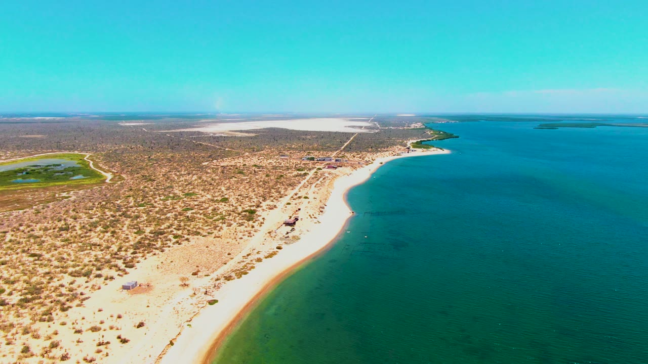 Opening aerial frame over San Carlos, Baja California Sur, showing a curved sandy beach between turquoise water and dry scrubland, with no visible people.