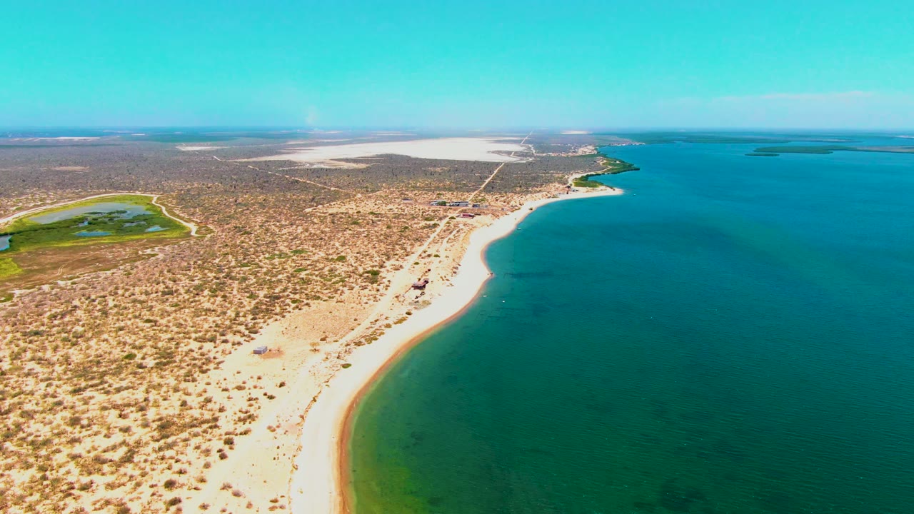 Aerial view in San Carlos, Baja California Sur with the beach curve closer to center, shallow green water near shore, deeper blue water offshore, dry vegetation inland, and no visible people.