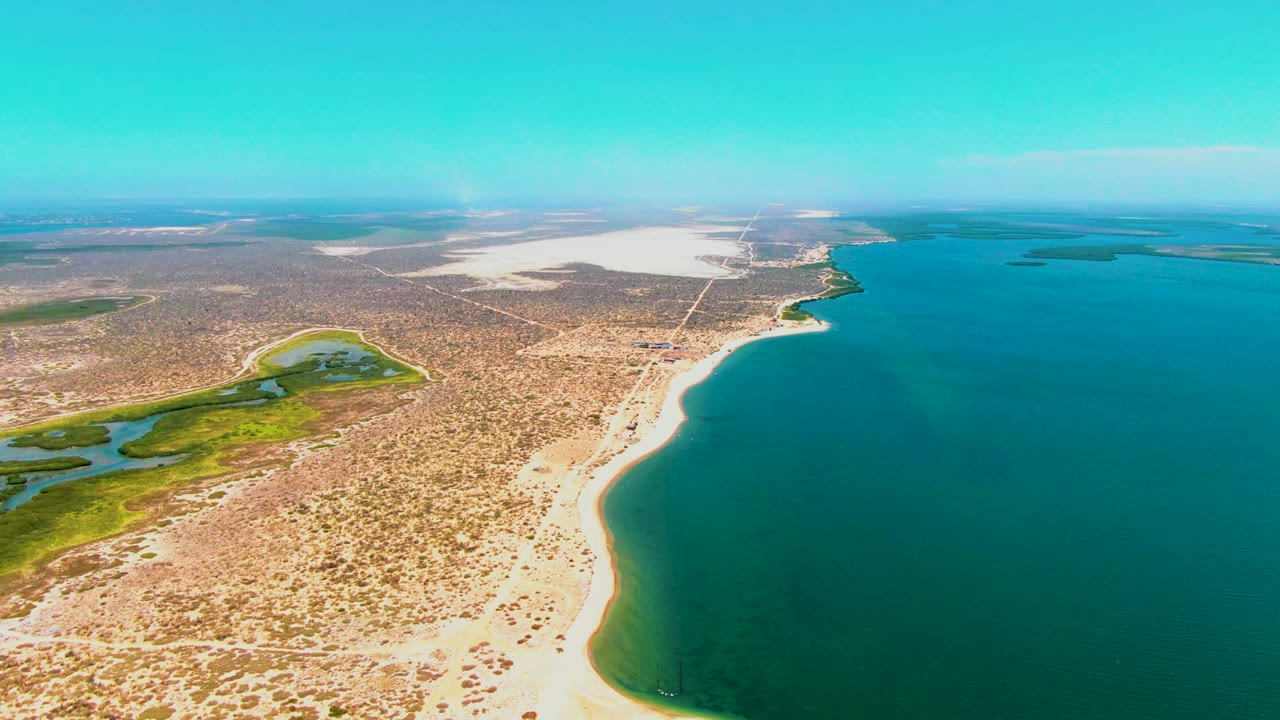 Aerial view of San Carlos, Baja California Sur, with the sandy shoreline arcing through the lower frame, turquoise sea to the right, dry patterned desert inland, and no visible people.