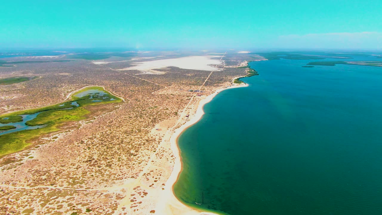 High aerial view over San Carlos, Baja California Sur, with a sandy beach curving beside turquoise ocean, scrub desert inland, a green wetland to the left, and no visible people.