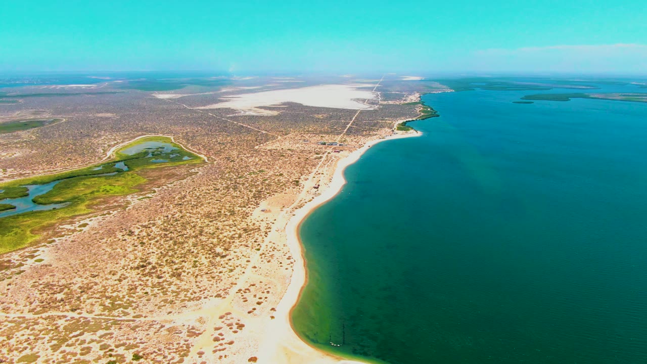 Oblique drone view over San Carlos, Baja California Sur, with the beach running diagonally through frame, darker water near shore, wetland greens inland, a straight road crossing the desert, and no visible people.