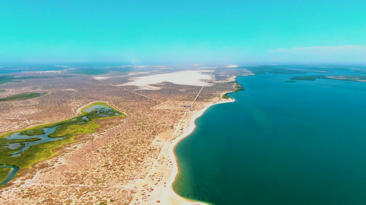 Opening aerial frame over San Carlos, Baja California Sur, with turquoise water on the right, a pale beach curving through dry scrubland, green wetland channels on the left, and no visible people.